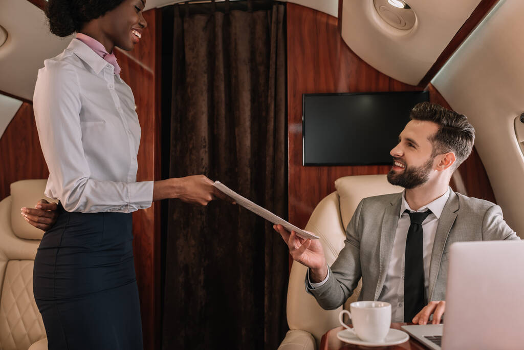 atractiva azafata afroamericana dando periódicos a hombre de negocios sonriente sentado en el portátil en jet privado
 - Foto, imagen