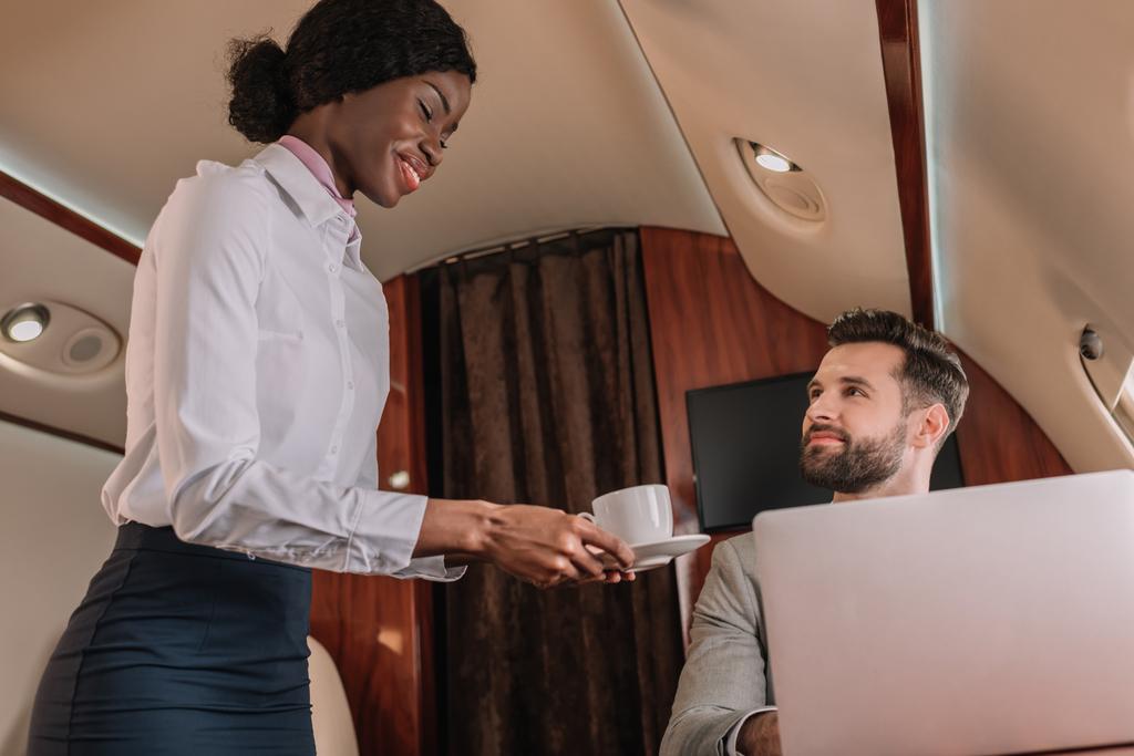 smiling african american stewardess giving cup of coffee to handsome businessman working on laptop in private jet - Photo, Image