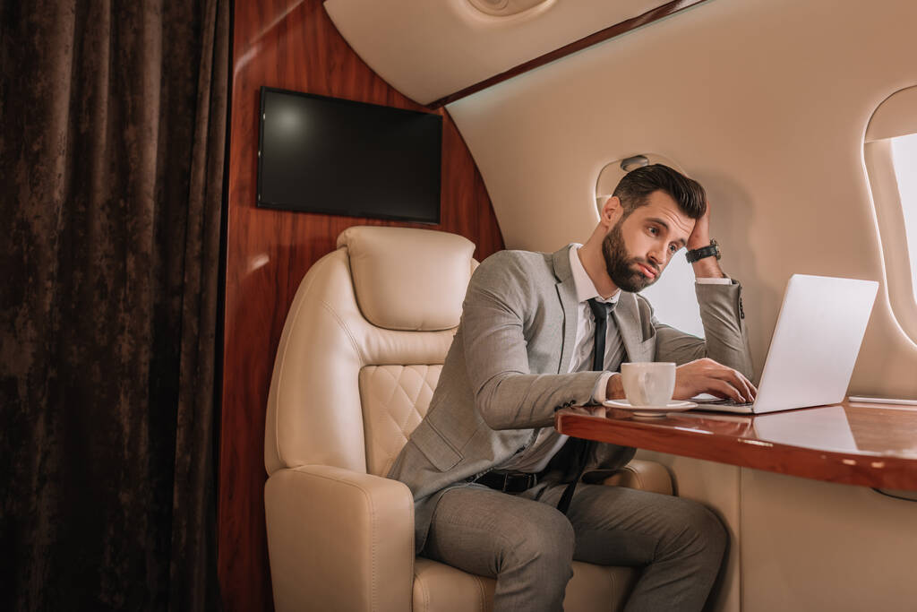 нудний бізнесмен торкається голови під час роботи на ноутбуці в приватному літаку
 - Фото, зображення