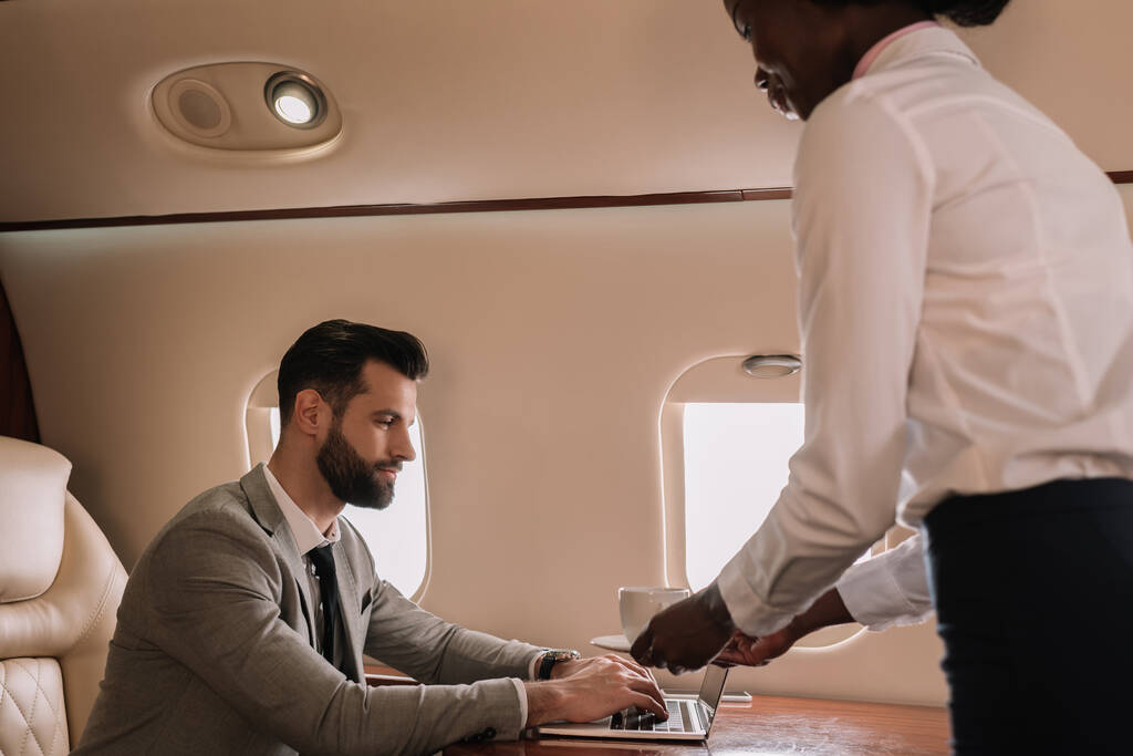 aeromoça americana africana dando xícara de café para homem de negócios trabalhando em laptop em avião privado
 - Foto, Imagem