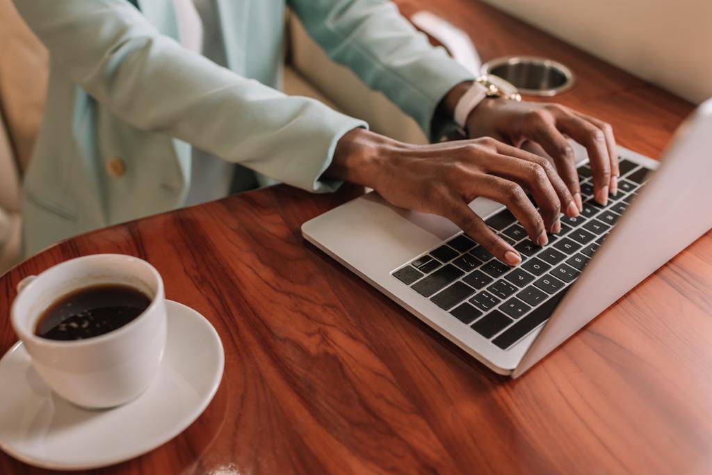 abgeschnittene Ansicht einer afrikanisch-amerikanischen Geschäftsfrau, die in der Nähe einer Tasse Kaffee auf ihrem Laptop tippt - Foto, Bild