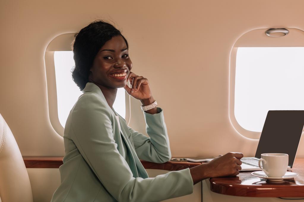 attraktive afrikanisch-amerikanische Geschäftsfrau lächelt in der Nähe von Laptop und Kaffeetasse im Privatjet in die Kamera - Foto, Bild