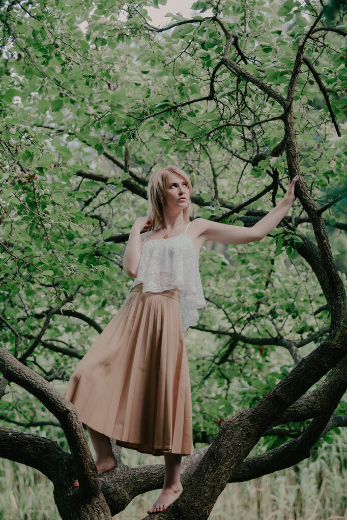 Портрет у природі. Струнка босоніжка в білій верхівці і спідниця стоїть на гілці дерева на літній да
 - Фото, зображення