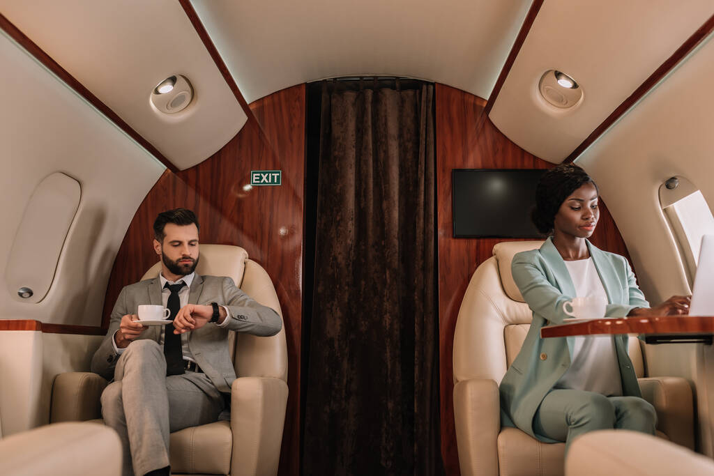 Aufmerksame afrikanisch-amerikanische Geschäftsfrau tippt auf Laptop, und Geschäftsmann schaut auf Armbanduhr, während er eine Tasse Kaffee in der Hand hält - Foto, Bild