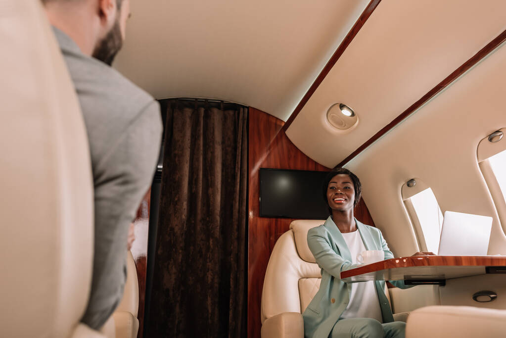 vista trasera del hombre de negocios mirando a la sonriente mujer afroamericana mientras viaja en avión
 - Foto, Imagen