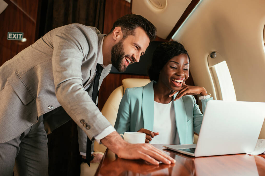 sonriente hombre de negocios de pie cerca de la feliz mujer de negocios afroamericana mientras que tiene videollamada en jet privado
 - Foto, Imagen