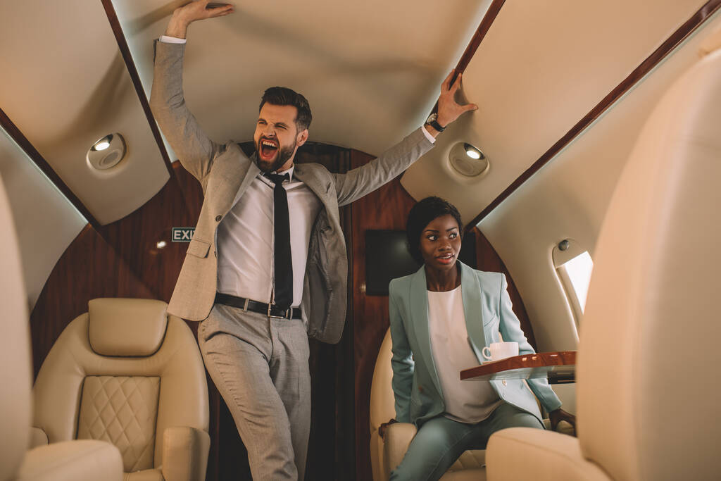 emocionado hombre de negocios gritando y tocando techo mientras asustada mujer de negocios afroamericana sentado en jet privado
 - Foto, Imagen