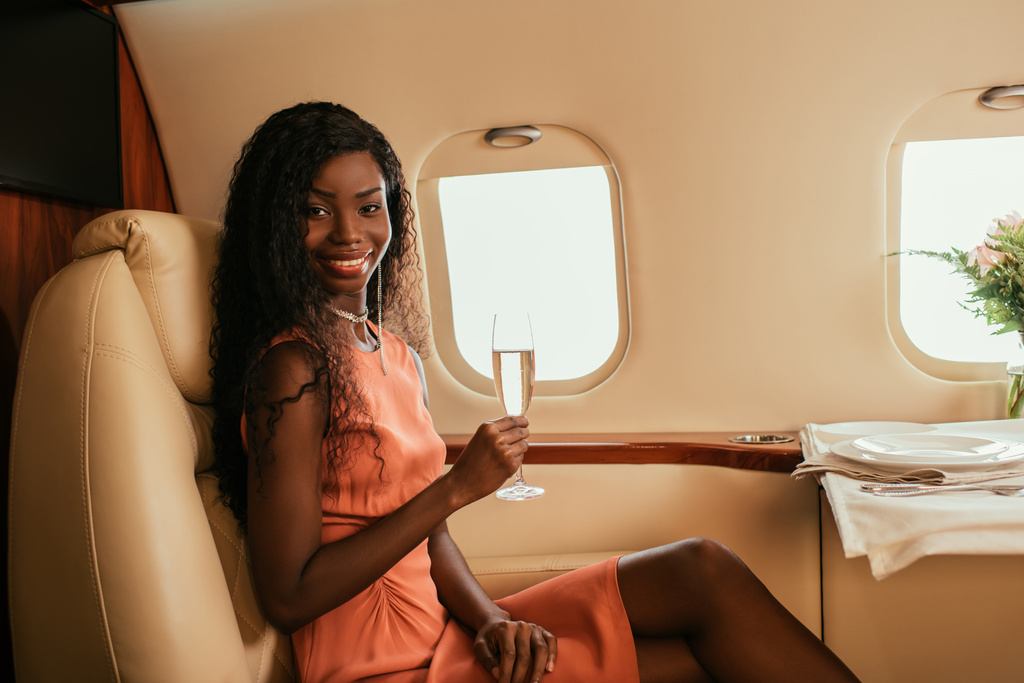 atractiva mujer afroamericana hoding copa de champán y sonriendo a la cámara mientras está sentado cerca de la mesa servida en avión privado
 - Foto, Imagen