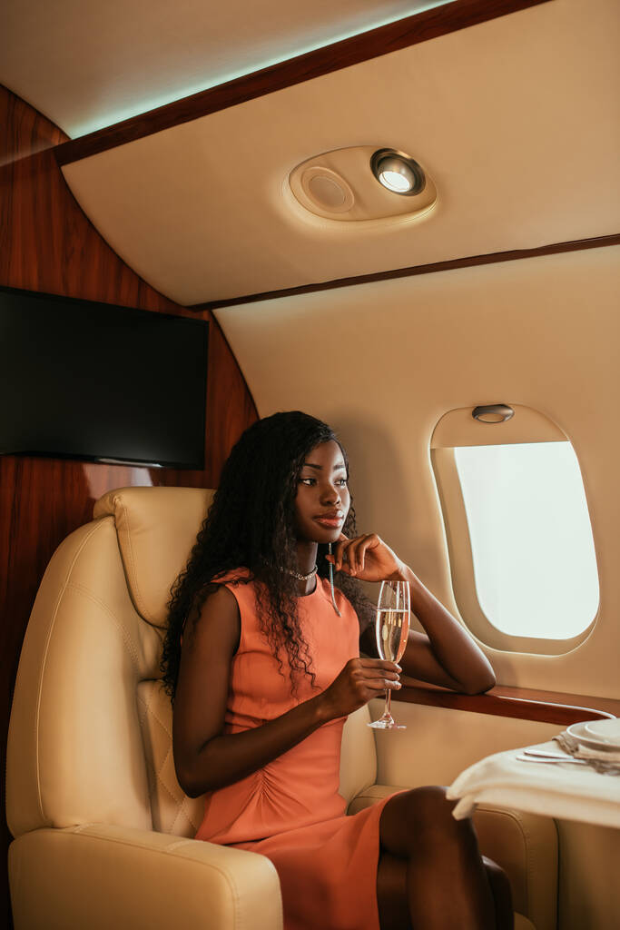 elegante, soñadora mujer afroamericana sosteniendo una copa de champán y mirando en un ojo de buey en un avión privado
 - Foto, Imagen