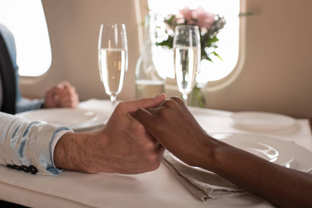 ostříhaný pohled na mezirasový pár držící se za ruce u sklenic šampaňského na servírovaném stole v soukromém tryskáči - Fotografie, Obrázek