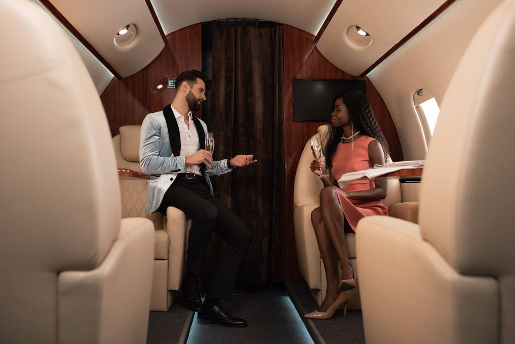 вибірковий фокус елегантної міжрасової пари, що розмовляє в приватному реактивному літаку, тримаючи шампанські окуляри
 - Фото, зображення