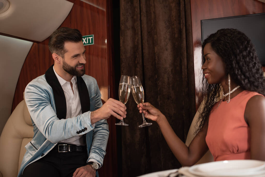 feliz, elegante pareja interracial tintineo copas de champán en avión privado
 - Foto, Imagen