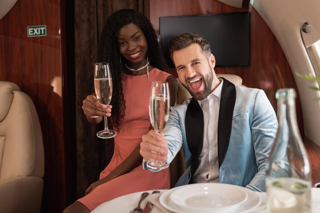 cheerful, elegant interracial couple holding glasses of champagne while looking at camera in private plane - Photo, Image