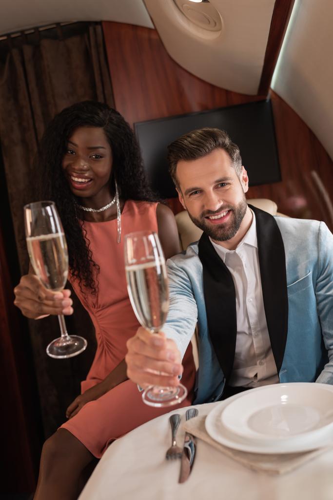 heureux, élégant couple interracial tenant des verres de champagne tout en regardant la caméra en jet privé
 - Photo, image