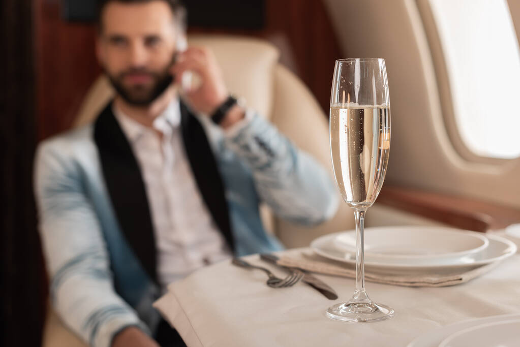 selective focus of elegant man talking on smartphone near glass of champagne in private jet - Photo, Image