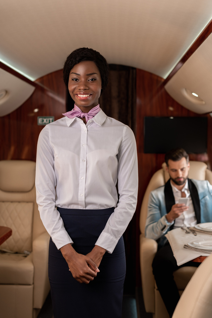 glimlachende Afrikaanse Amerikaanse stewardess op zoek naar camera, terwijl elegante man met behulp van smartphone in prive-vliegtuig - Foto, afbeelding