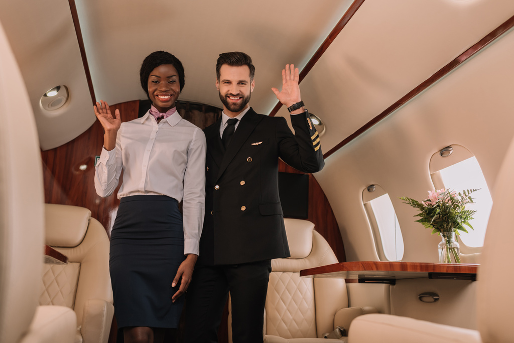 Mutlu pilot ve gülümseyen Afro-Amerikan hostes özel jette kameraya bakarken el sallıyor. - Fotoğraf, Görsel