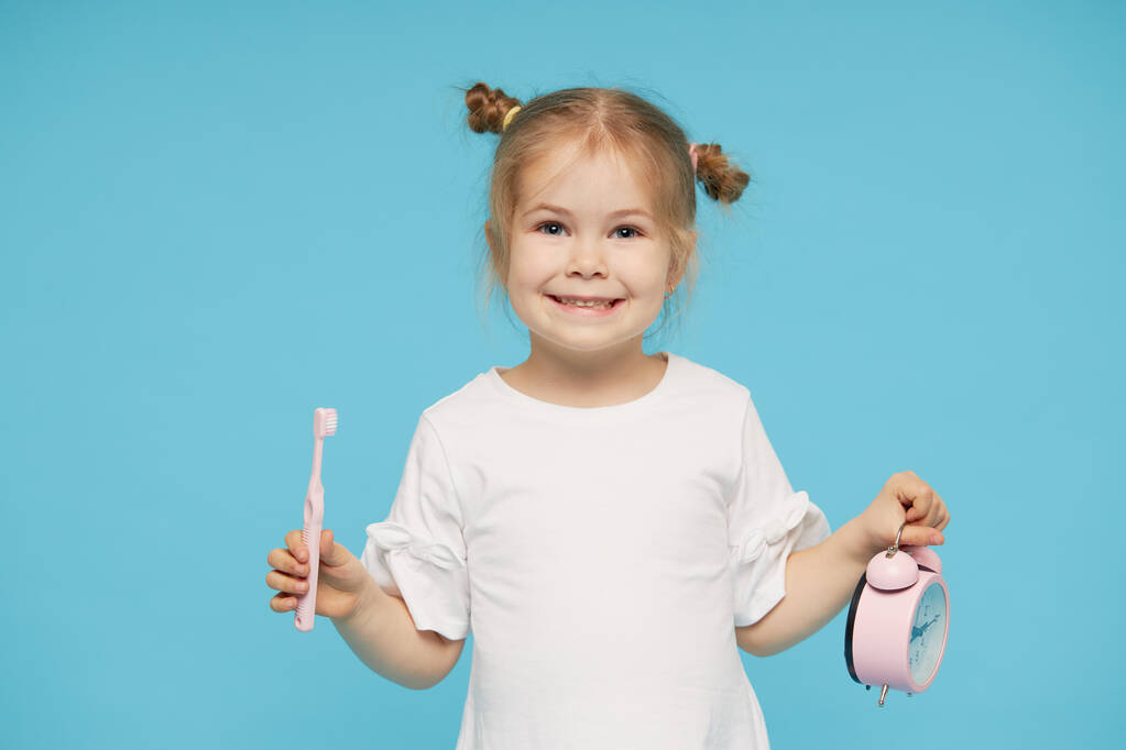 Suuhygienia. Aika harjata hampaat. Pieni lapsi tyttö harjaamalla hampaitaan hammasharjalla ja katsomalla kameraa sinisellä taustalla
 - Valokuva, kuva