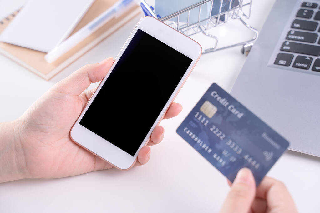 Frau mit Smartphone, Konzept des Online-Bezahlens zu Hause bleiben, elektronisches Bezahlen auf weißem Tischhintergrund mit Einkaufswagen, Nahaufnahme. - Foto, Bild