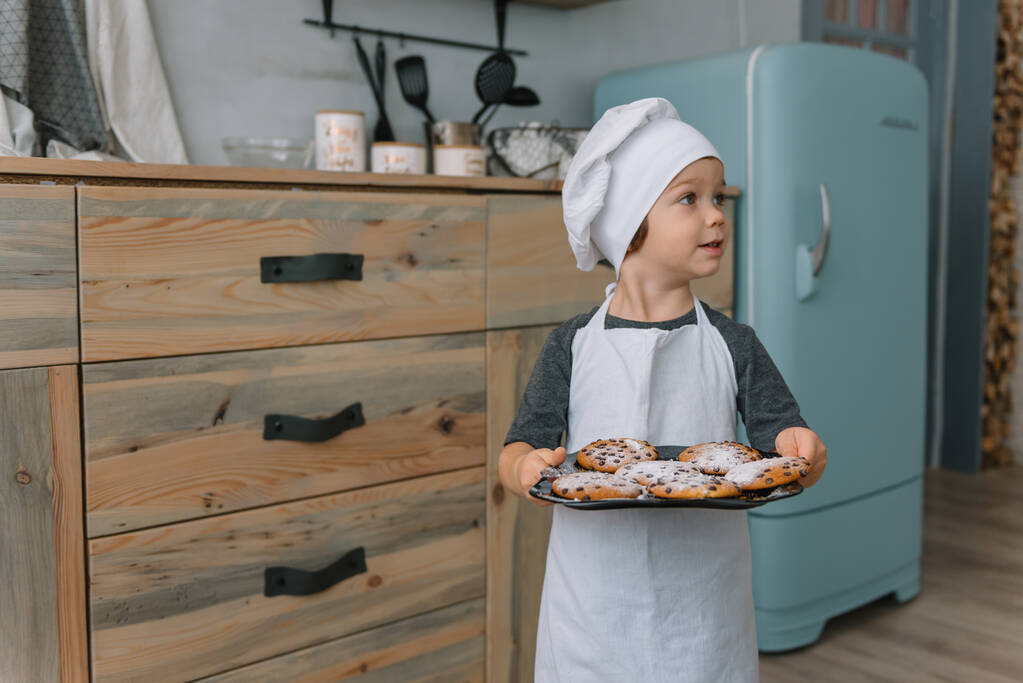 Genç mutlu anne ve bebeği mutfakta kurabiye pişiriyor. Noel ev yapımı zencefilli çörek. Beyaz üniformalı, şapkalı, çikolatalı kurabiyeli tatlı çocuk. - Fotoğraf, Görsel