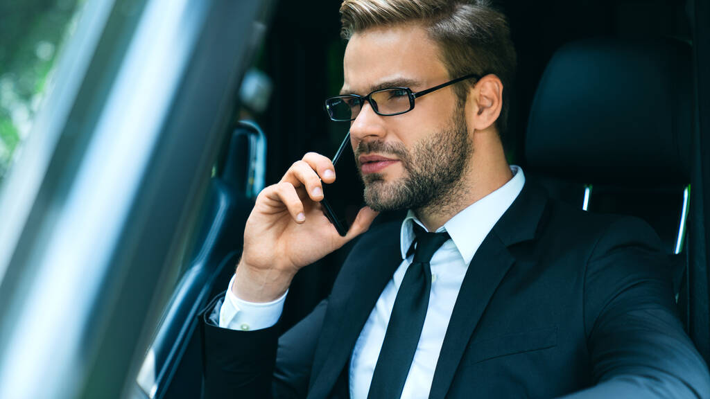 Jóképű fiatal üzletember teljes öltönyben beszél az okostelefonon vezetés közben. - Fotó, kép