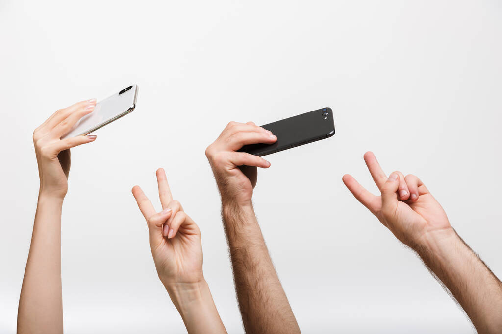 Κοντινή εικόνα των χεριών ενός άνδρα και της γυναίκας που απομονώνονται πάνω από το λευκό φόντο τοίχο κρατώντας κινητά τηλέφωνα λάβει μια selfie δείχνει χειρονομία ειρήνης. - Φωτογραφία, εικόνα