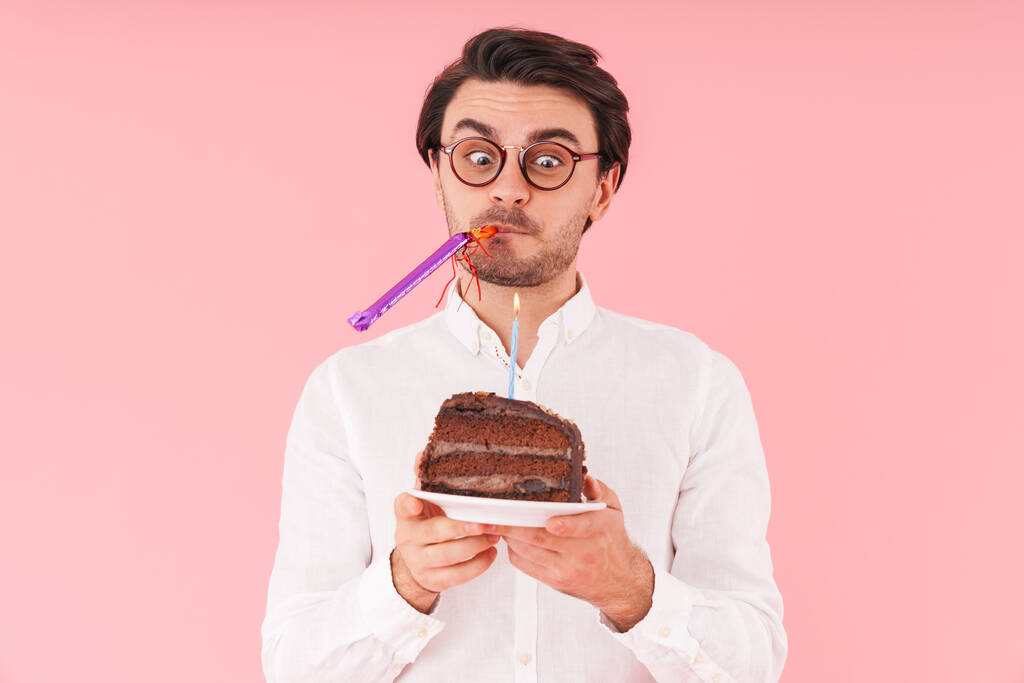 Immagine di un giovane uomo sorpreso con gli occhiali in mano torta al cioccolato con candela isolata su sfondo rosa
 - Foto, immagini