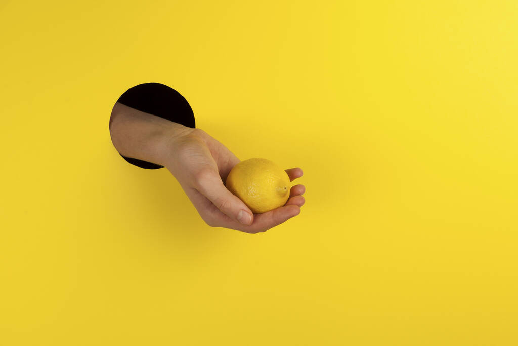 黄色い背景の穴に手を取り、ビタミンCの源としてレモンを提供し、風邪やウイルス性疾患の予防。スペースのコピー. - 写真・画像