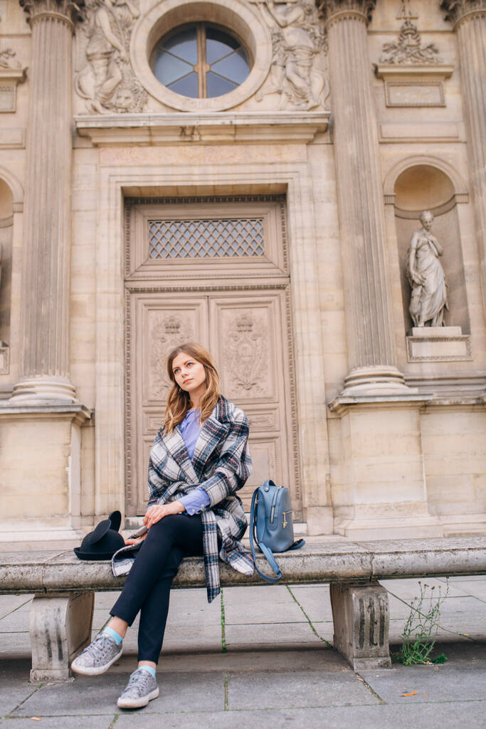 Tyttö kauniissa takissa istuu penkillä Louvren lähellä. Käsite vierailevat Ranskassa ja historiallisia rakennuksia
. - Valokuva, kuva