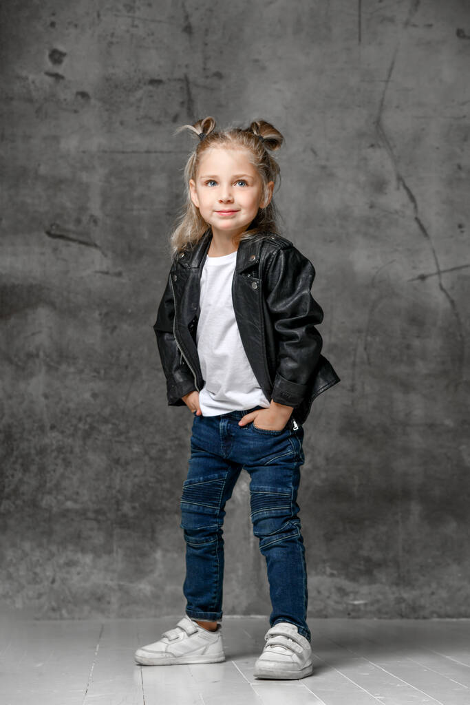 Pequena menina loira sorridente em estilo rock elegante jaqueta de couro preto, jeans azuis e tênis brancos de pé com as mãos nos bolsos
 - Foto, Imagem
