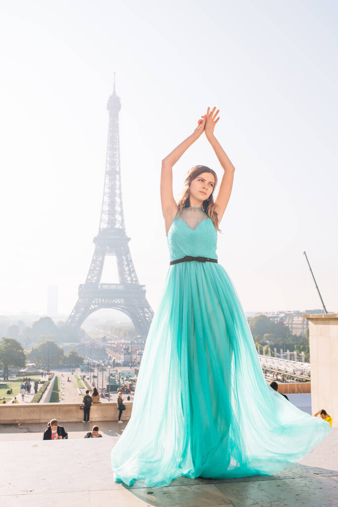 Ein junges schönes und elegantes Mädchen in einem blau-grünen Kleid steht vor dem Hintergrund des Eiffelturms in Paris. - Foto, Bild