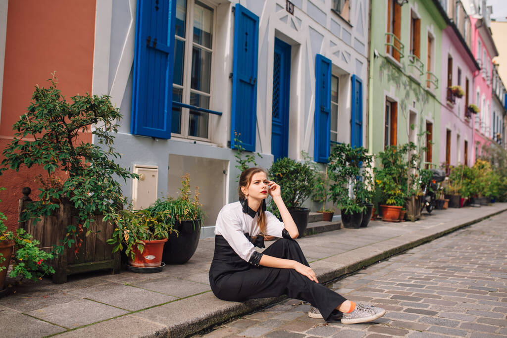 Une belle jeune femme en combinaison noire à la mode est assise sur un porche dans une rue parisienne avec des maisons colorées et lumineuses.France
 - Photo, image