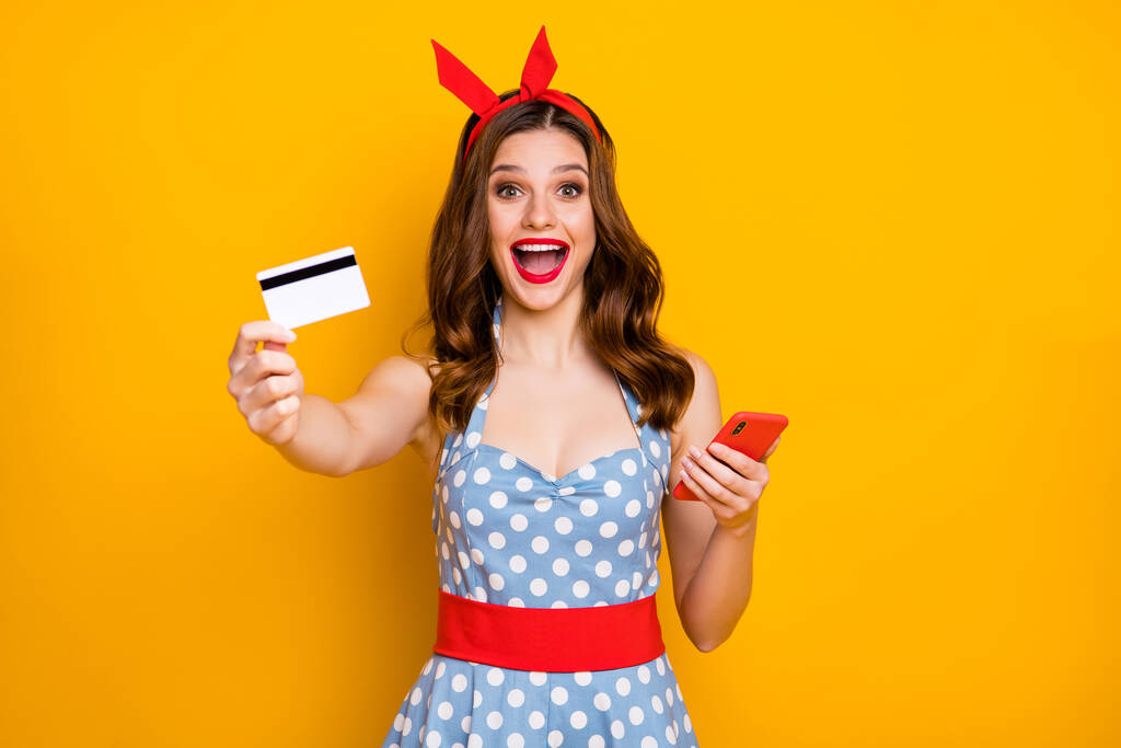 Őrült sokkos lány lenyűgözött egyszerű internetes fizetés mobiltelefon ő tartja show műanyag kártya élvezze kedvezmények viselni kék pontozott ruha piros évjárat fejpánt elszigetelt élénk sárga háttér - Fotó, kép