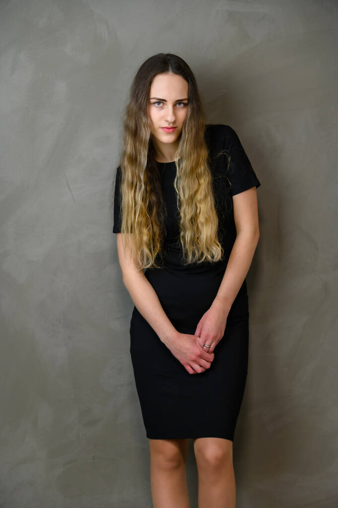 Вертикальне фото милої білошкірої дівчини-підлітка в чорній сукні з довгим волоссям в студії на сірому фоні
 - Фото, зображення