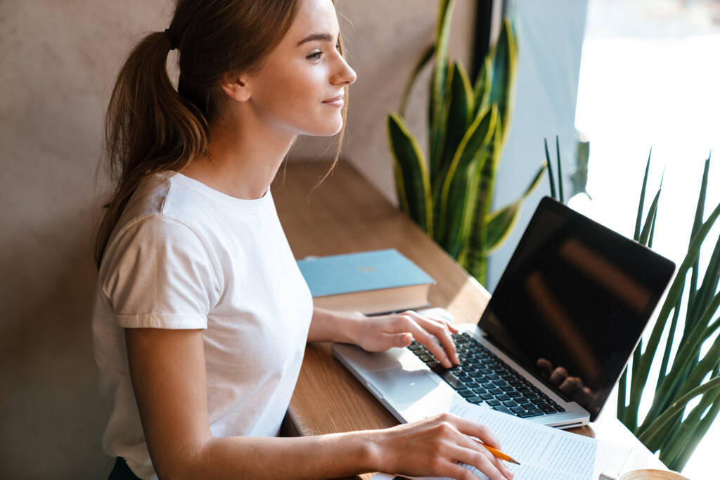 Photo de femme mignonne heureuse utilisant un ordinateur portable et étudiant avec un cahier d'exercices tout en étant assise dans un café confortable
 - Photo, image