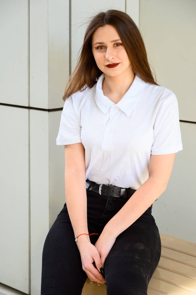 Retrato de uma jovem morena caucasiana em uma camiseta branca ao ar livre em um dia ensolarado na cidade contra o fundo de uma parede do edifício
. - Foto, Imagem