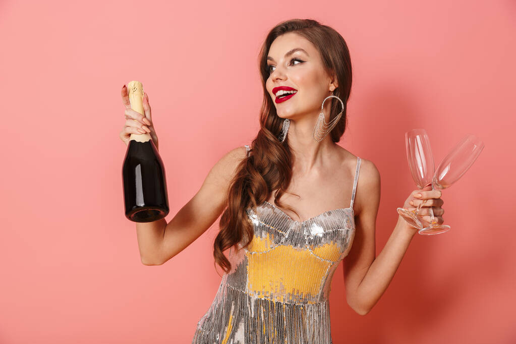 Фото молодой веселой позитивной женщины в ярких блестках платье изолированы на розовом фоне стены держа бутылку шампанского и очки
. - Фото, изображение