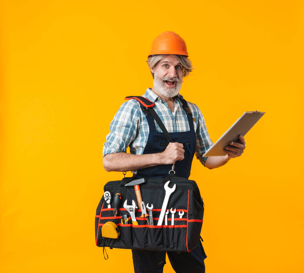Obrázek překvapené starší šedovlasý vousatý muž stavitel v helmě pózování izolované přes žlutou stěnu pozadí podpírání taška s vybavením nástroje a schránky. - Fotografie, Obrázek