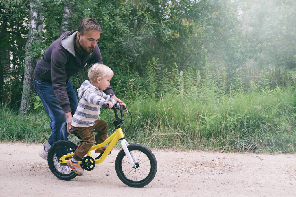Vader leert zijn kleine kind fietsen in het voorjaarspark. Gelukkige familiemomenten. Samen tijd doorbrengen met vader en zoon. Candid lifestyle imago. - Foto, afbeelding