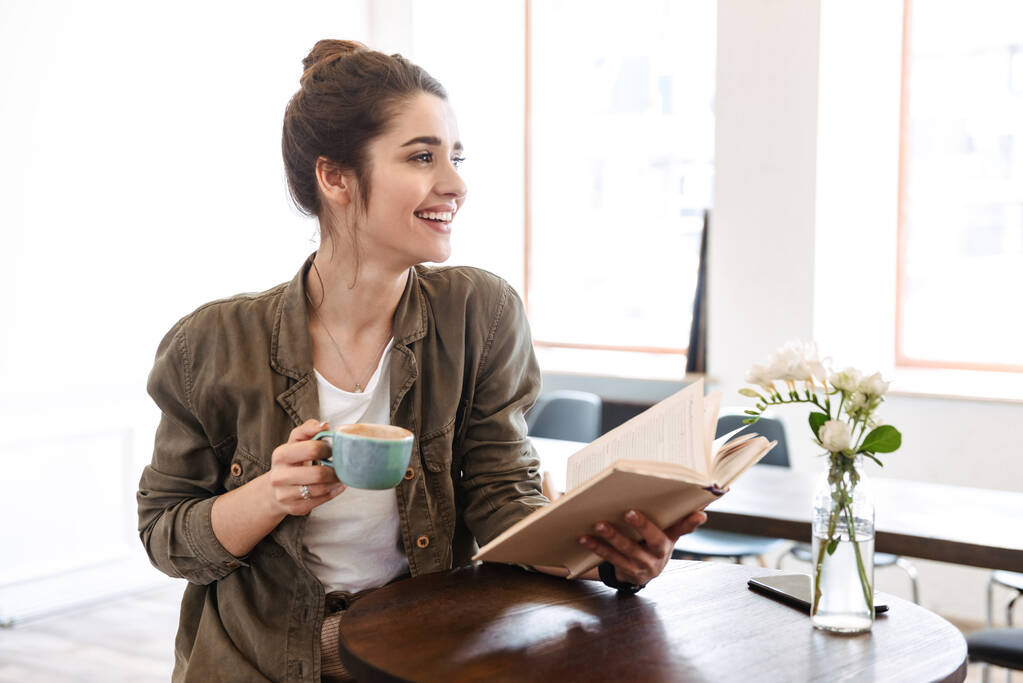 Bella giovane donna che legge il libro mentre si siede al caffè in casa a bere caffè
 - Foto, immagini