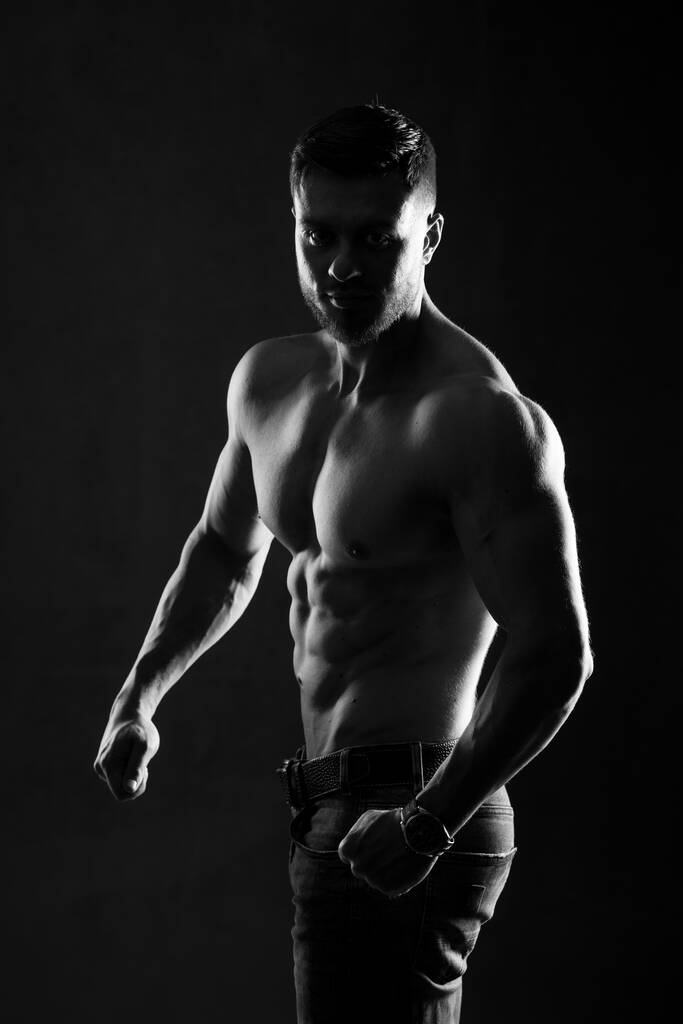 Сексуальный мускулистый мужчина позирует на тёмном фоне с голым туловищем. Рука об руку. Мышечное тело и сильный пресс. Черное и белое. Снимок студии
. - Фото, изображение