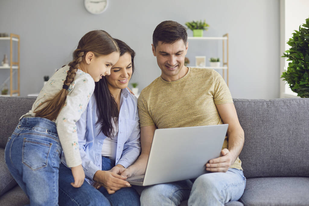 Szczęśliwa rodzina na czacie online za pomocą laptopa podczas siedzenia z dzieckiem na kanapie w domu. - Zdjęcie, obraz