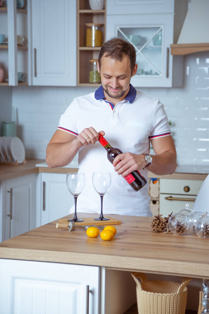 Genç adam mutfakta duruyor ve bardaklara kırmızı şarap dolduruyor. - Fotoğraf, Görsel
