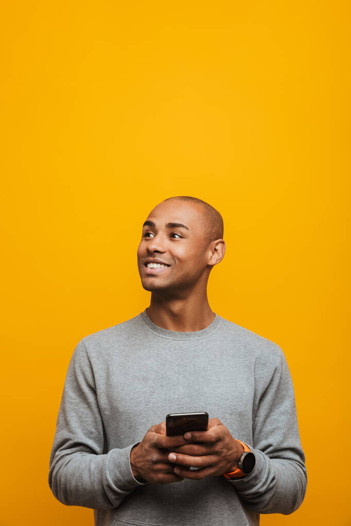 Πορτρέτο ενός ελκυστικού χαμογελαστού ανέμελου νεαρού αφρικανού άνδρα που στέκεται πάνω από κίτρινο φόντο, χρησιμοποιώντας το κινητό τηλέφωνο, κοιτάζοντας μακριά - Φωτογραφία, εικόνα