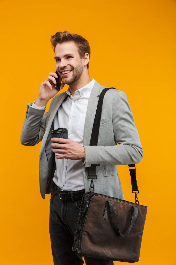 Przystojny uśmiechnięty młody biznesmen w kurtce stojący na żółtym tle, niosący torbę, rozmawiający przez telefon komórkowy - Zdjęcie, obraz