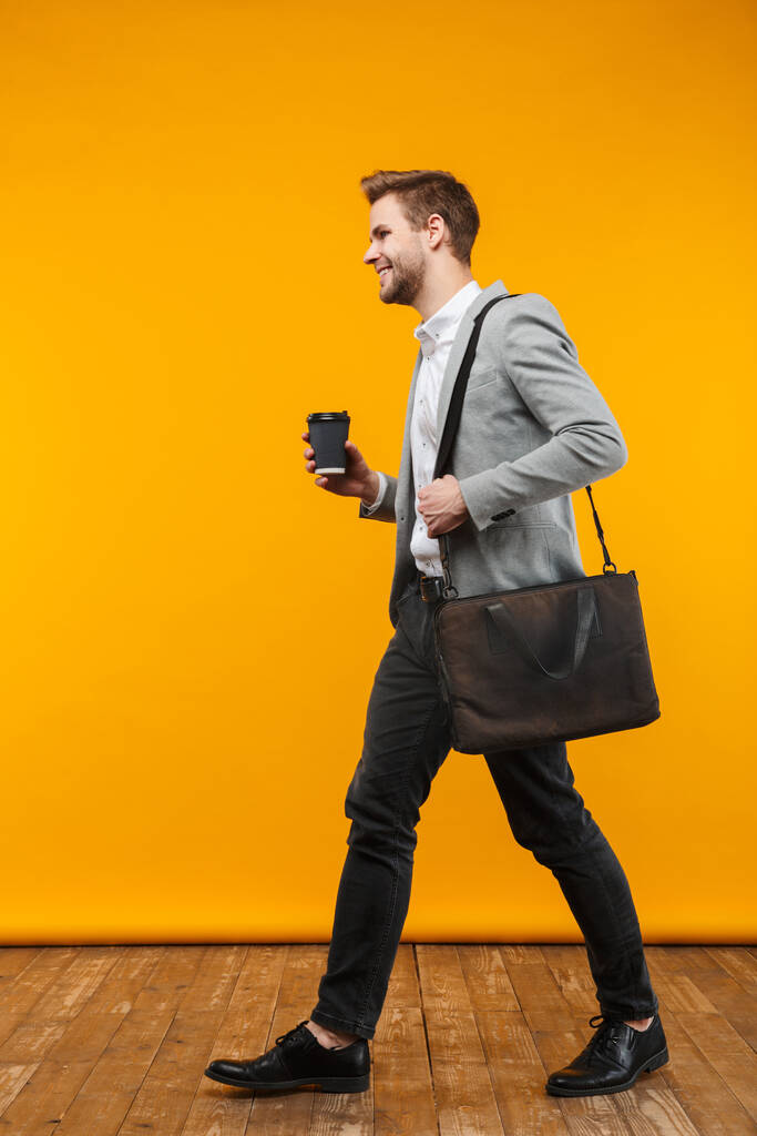 Comprimento total de um belo sorridente jovem empresário vestindo jaqueta andando sobre fundo amarelo, segurando taça de café takeaway
 - Foto, Imagem