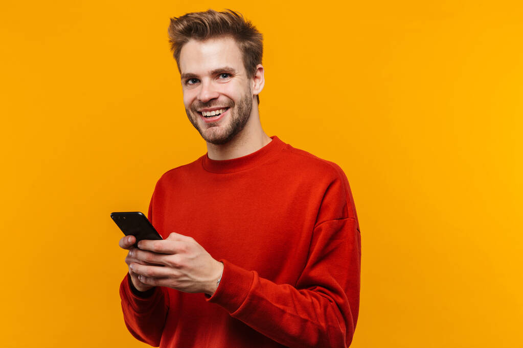 Attraktiver, glücklicher junger Mann mit Pullover, der isoliert vor gelbem Hintergrund steht und Mobiltelefone benutzt - Foto, Bild