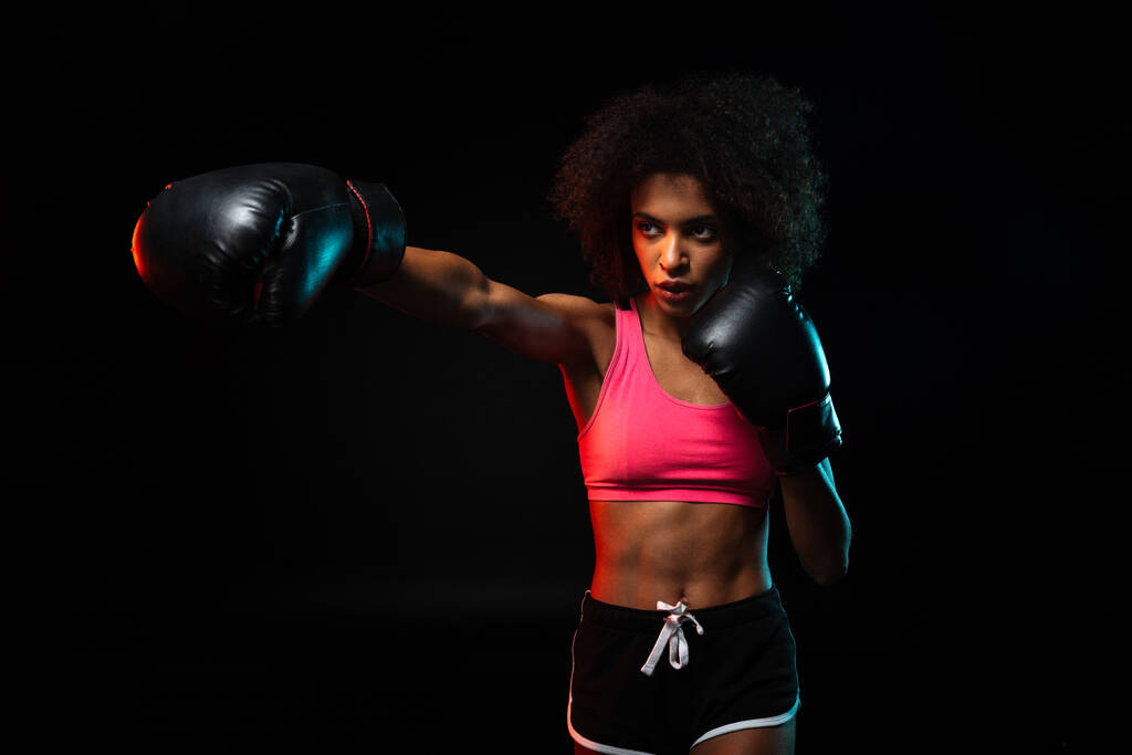 Image of young african american sportswoman working out in boxing gloves isolated over black background - Photo, Image