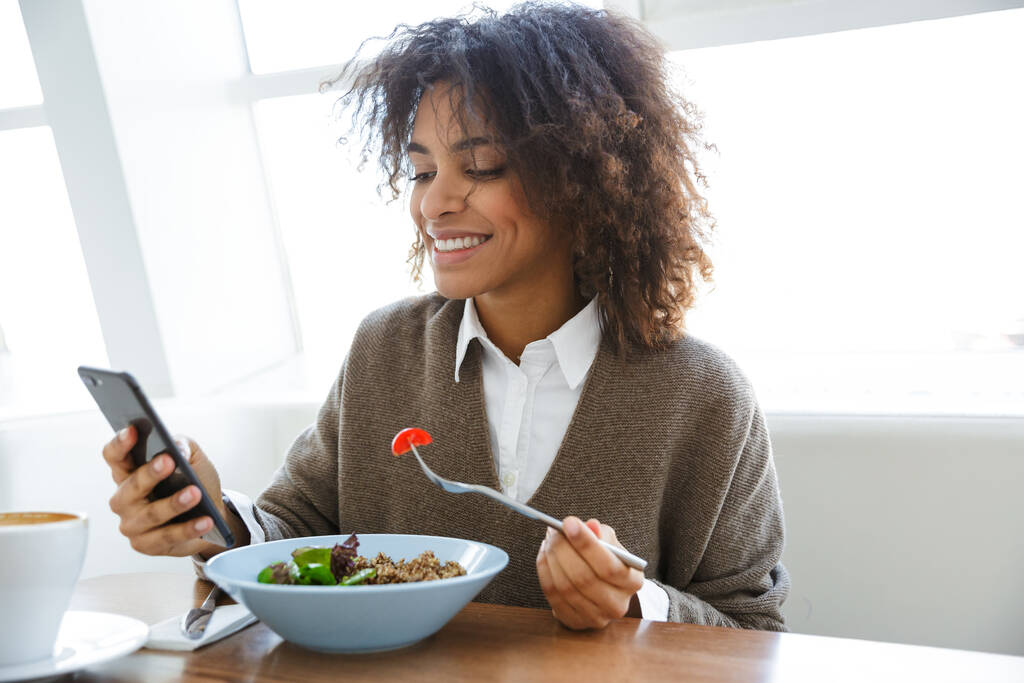 Πορτρέτο της νεαρής όμορφης αφροαμερικάνας γυναίκας που χρησιμοποιεί κινητό τηλέφωνο ενώ γευματίζει στο καφέ - Φωτογραφία, εικόνα