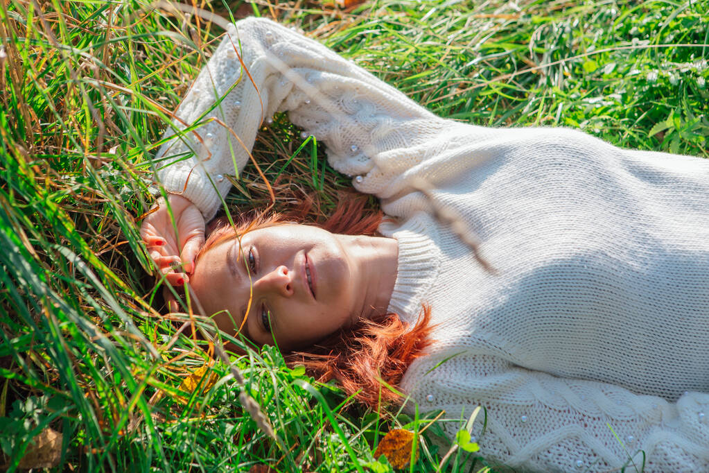 Portrait matinal d'une belle femme heureuse aux cheveux roux posée dans l'herbe en pull blanc
 - Photo, image